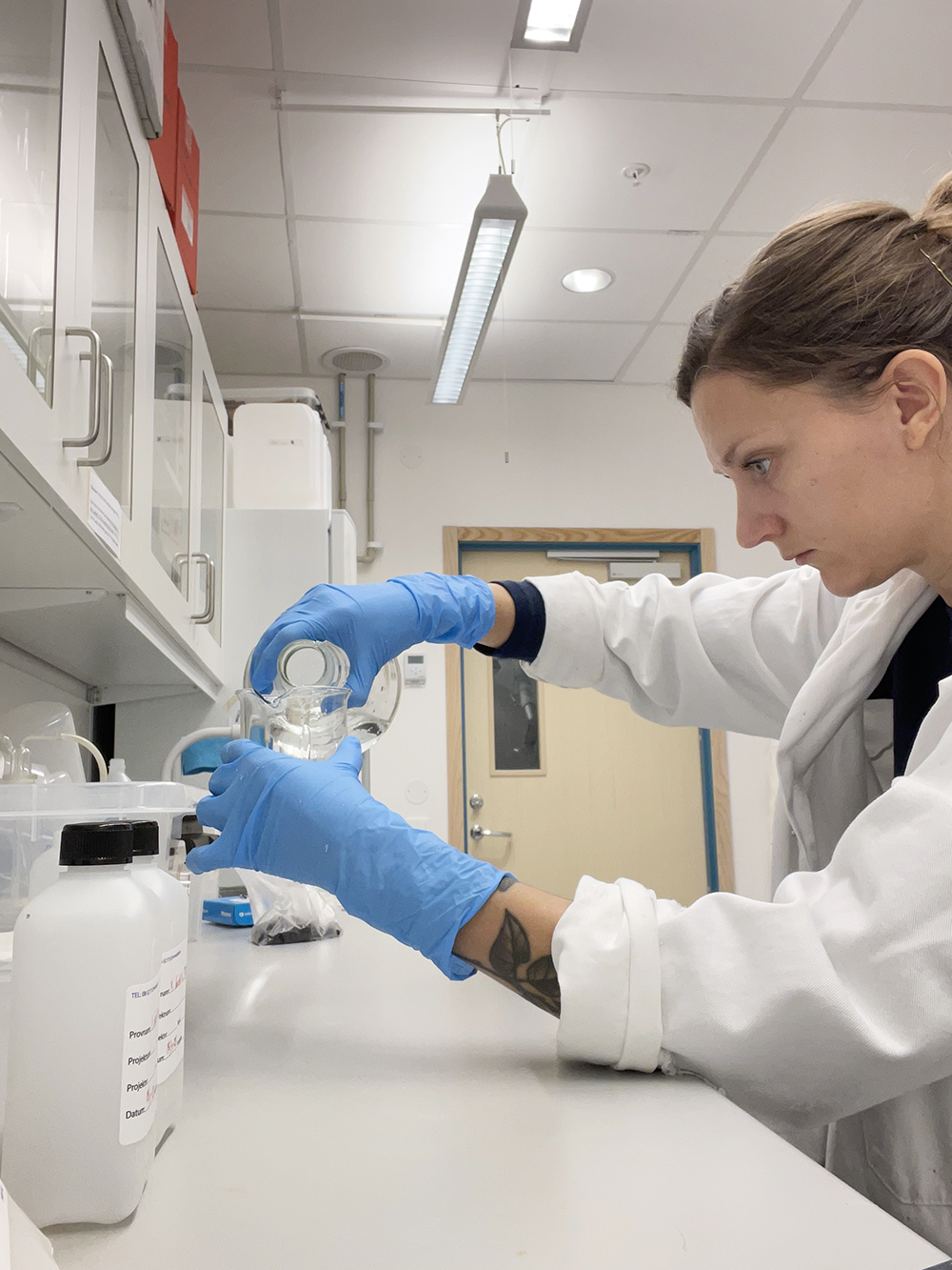 Kvinna i profil med labbrock och handskar fyller fokuserat en flaska med vätska i labb