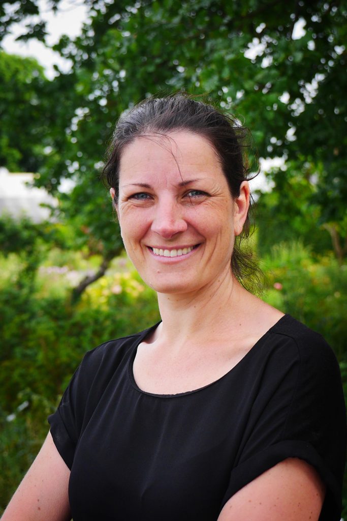 Ett porträtt på Lotta leendes utomhus en solig dag med grönska i bakgrunden