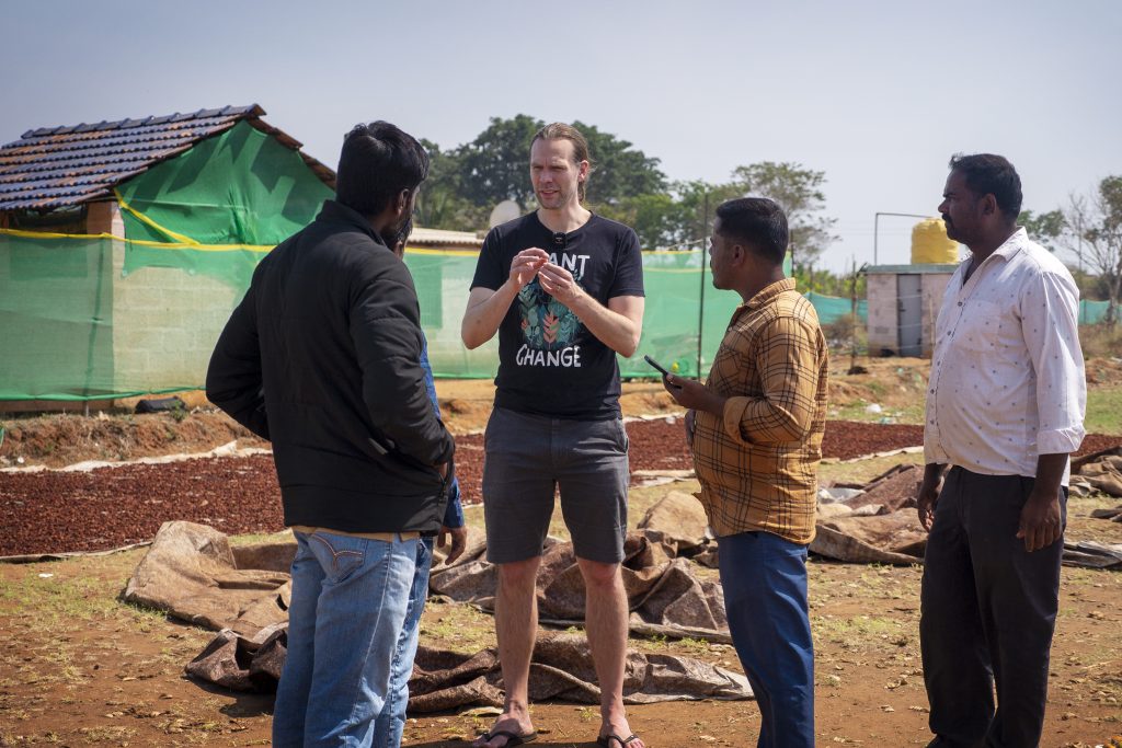 Mattias talar med Indier utomhus en solig dag och i bakgrunden skymtas nötter som ligger på tork.
